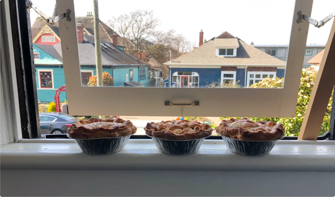 Three pies cooling on a windowsill with colorful houses in the background.